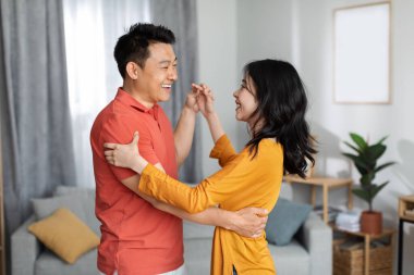 Beautiful asian couple enjoying time together at home, dancing waltz, happy chinese middle aged man and pretty young woman in homewear holding hands and smiling to each other, copy space