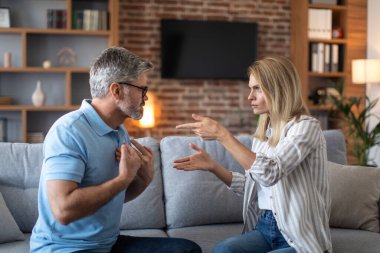 Upset offended angry excited mature caucasian husband and wife quarreling in living room interior, profile. Couple suffering from stress, conflict, relationship problems, scandal and negative emotions