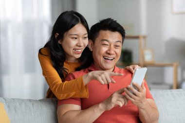 Happy japanese couple using brand new smartphone together at home, cheerful middle aged man and young woman watching photos on mobile phone and smiling, using mobile app, copy space