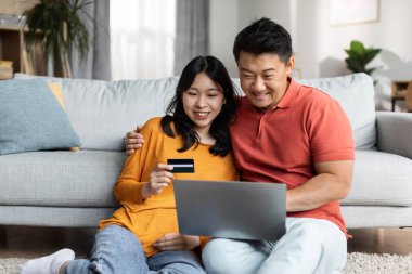Relaxed japanese spouses sitting on floor by couch, using notebook and credit card, looking at laptop screen and smiling, asian husband and wife shopping on Internet, copy space