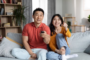 Happy asian middle aged man and young woman in homewear sitting on sofa in living room, watching TV together at home, holding remote and pointing at camera, copy space