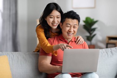 Loving chinese spouses shopping on Internet from home, cheerful young asian woman hugging her husband from behind, pointing at laptop screen, family choosing new furniture online, copy space
