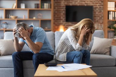 Unhappy mature caucasian couple holding heads with hands, suffer from debts in room interior with documents and calculator. Taxes, problems with bills, home bookkeeping, credit, bankruptcy and stress
