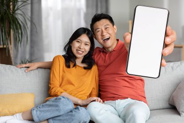 Cheerful chinese lovers man and woman in homewear sitting on couch, embracing and showing cellphone with white empty screen, recommending newest mobile app, home interior, copy space, mockup