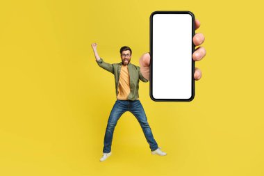 Full body length portrait of excited man jumping, making winner gesture with clenched fist and showing cellphone with empty space for mobile app or website design, yellow studio background