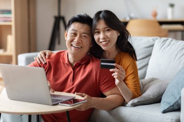 Happy asian middle aged husband and millennial wife sitting next to couch in living room, using computer and showing credit card, planning vacation, booking hotel and tickets online, copy space