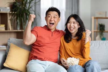 Emotional asian middle aged man and young woman in casual outfits sitting on couch, eating popcorn and raising hands up, celebrating victory, watching TV show or football game at home, copy space