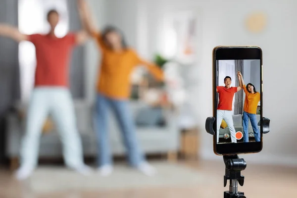 Funny asian husband and wife recording video for social media or personal family vlog, holding hands and dancing in front of camera, selective focus on modern cell phone on tripod, home interior