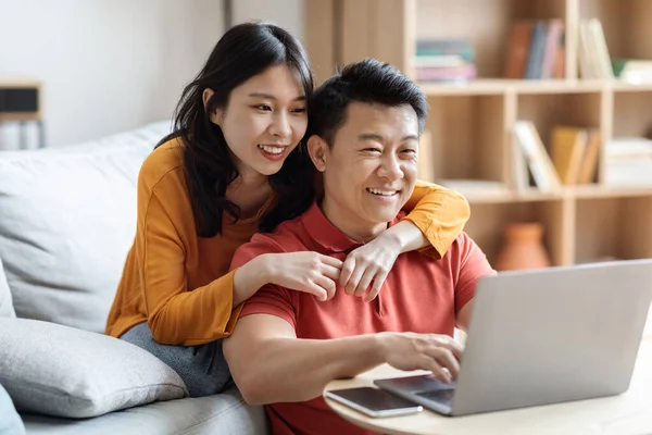 Happy japanese lovers planning vacation, cheerful asian middle eastern man and young woman sitting in living room, using computer, booking tickets and hotel on Internet, copy space