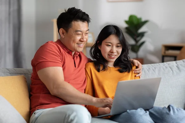 Asian spouses watching movie on Internet, using laptop at home. Smiling chinese man and woman sitting on couch, embracing, looking at computer screen, websurfing together, copy space