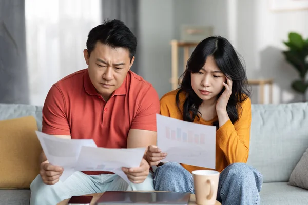 Confused asian couple checking bills, sitting on couch, reading documents, unhappy chinese man and woman holding papers, counting monthly spendings, copy space. Family budget concept