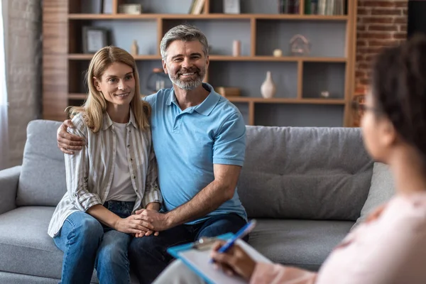 Smiling mature caucasian man and lady hugs at meeting with psychologist in office clinic interior with young african american doctor. Reconciliation, medical help and family therapy, problem solving