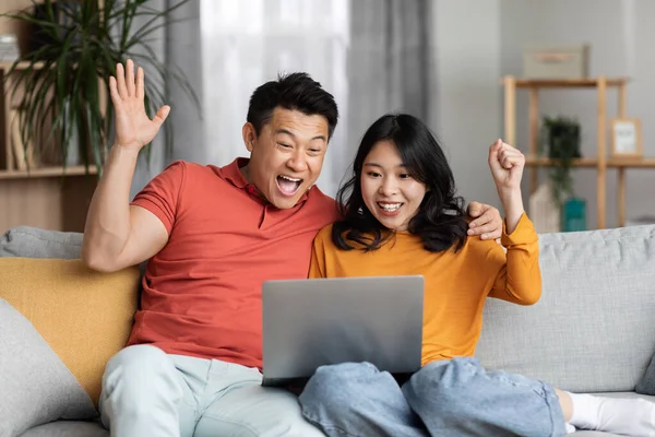 Emotional Asian Couple Gambling Internet Sitting Couch Home Raising Hands — Stock fotografie