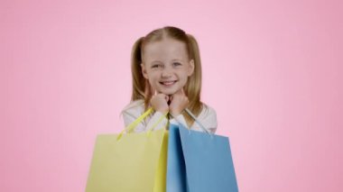 Joy of sales. Studio portrait of cute little girl shopaholic enjoying success, carrying blue and yellow shopping bags, pink background, slow motion