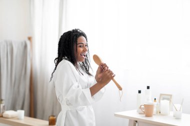 Morning Fun. Joyful African American Female Singing In Bathroom Interior, Cheerful Young Black Woman In White Silk Robe Using Body Brush As Microphone, Enjoying Home Beauty Routine, Copy Space