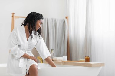 Bath Time. Beautiful Young Black Lady In White Silk Robe Sitting On Bathtub And Checking Water Temperature, Smiling African American Woman Relaxing In Modern Bathroom Interior At Home, Copy Space