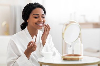 Attractive Black Female Looking At Mirror And Applying Moisturising Lip Balm, Beautiful African American Woman Holding Chapstick, Using Hygienical Lipstick While Sitting At Dressing Table At Home