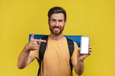 Online travel booking. Positive middle aged male tourist showing cellphone with empty screen and pointing on it over yellow studio background. Mockup for your design