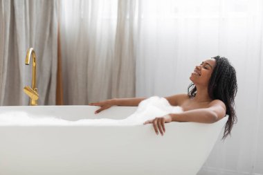 Domestic Spa. Beautiful black woman relaxing in bathtub filled with foam, smiling african american enjoying hot bath with soap bubbles, resting in modern bathroom interior, side view with copy space
