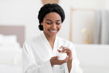 Skincare Cosmetics. Smiling black female trying new moisturising cream while making beauty routine at home, happy african american woman holding open jar with body lotion, enjoying selfcare, closeup