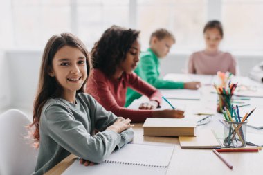 Happy Schoolgirl Sitting At Desk Smiling To Camera Learning And Posing With Multicultural School Kids In Classroom Indoor. Modern Education, Back To School Concept. Selective Focus