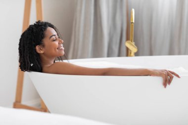 Bodycare Concept. Attractive Black Woman Lying In Bathtub In Luxury Bathroom Interior, Smiling Beautiful African American Female Taking Hot Bath At Home, Enjoying Domestic Spa, Side View
