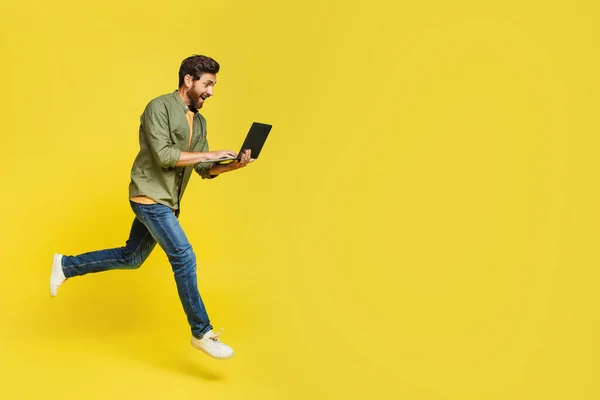 Full length of excited middle aged man using laptop computer and jumping on yellow background copy space. Stylish freelancer male flying in air with modern portable pc