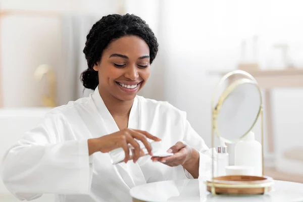 Smiling Black Woman Pouring Micellar Water On Cotton Pad While Making Beauty Routine At Home, Attractive Young African American Female Sitting Near Mirror, Cleansing Face From Makeup, Closeup