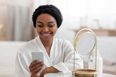Happy Beautiful Black Female Usign Smartphone While Getting Ready Near Mirror At Home, Young African American Woman Wearing White Silk Robe Messaging On Mobile Phone Or Making Online Shopping