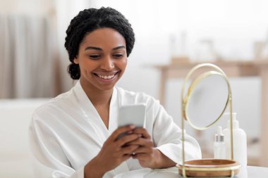 Smiling Black Woman With Smartphone Reading Beauty Hacks While Getting Ready Near Mirror At Home, Attractive African American Female Wearing White Silk Robe Browsing Internet On Mobile Phone