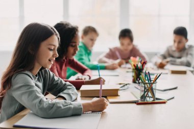 Back To School. Schoolgirl Writing Essay During Class Sitting With Mixed Children In Modern Classroom. Girl Taking Notes Learning Indoor. Knowledge And Education. Selective Focus