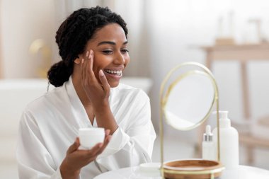 Anti-Aging Skincare. Portrait Of Smiling Attractive Black Female Applying Moisturising Face Cream While Sitting In Front Of Mirror At Home, African American Female Enjoying Beauty Routine, Free Space