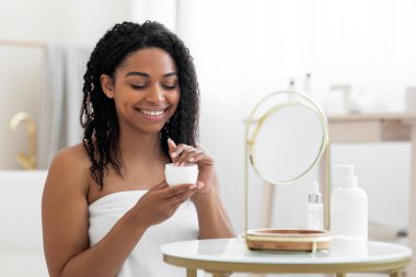 Smiling black woman taking moisturising cream from jar while making beauty treatments at home, attractive african american lady sitting wrapped in towel at dressing table, using new skincare cosmetic