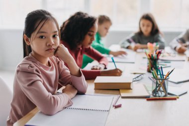 Sad Asian Schoolgirl Looking At Camera Sitting At Desk With Multiethnic Classmates During Class In Modern Classroom Indoor. Child And School Education Concept. Selective Focus