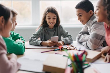 Cheerful Diverse School Children Using Cellphone Sitting At Desk In Modern Classroom. Classmates Using Educational Application On Smartphone Together And Having Fun At School. Kids And Gadgets