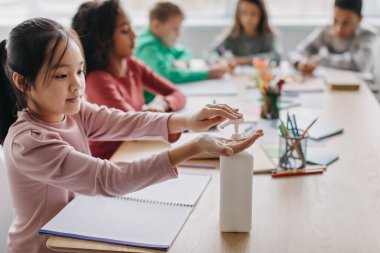 Covid 19 Prevention. Japanese Schoolgirl Disinfecting Hands At School. Kid Girl Using Sanitizer During Class Sitting With Diverse Classmates In Modern Classroom Indoors. Children Healthcare Concept