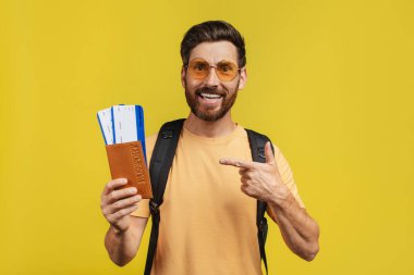 Positive middle aged man with backpack pointing at passport and airplane tickets, posing over yellow studio background and smiling at camera