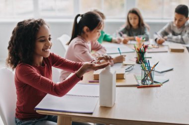 African American Schoolgirl Disinfecting Hands Applying Sanitizer Sitting With Multiethnic Classmates In Modern Classroom. School And Healthcare, Coronavirus Protection Concept