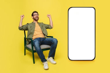 Great offer. Overjoyed man celebrating success near big smartphone with blank screen, clencing fists while sitting in armchair over yellow background, mockup