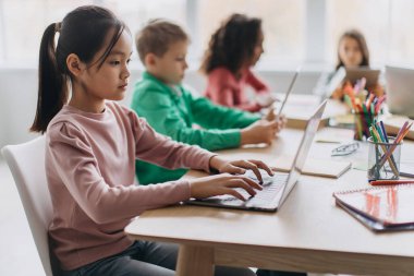 Korean Little Girl Using Laptop Sitting At Desk With Multiracial Classmates In Modern Classroom Indoor. E-Learning Website And Modern Internet Education Service. Selective Focus