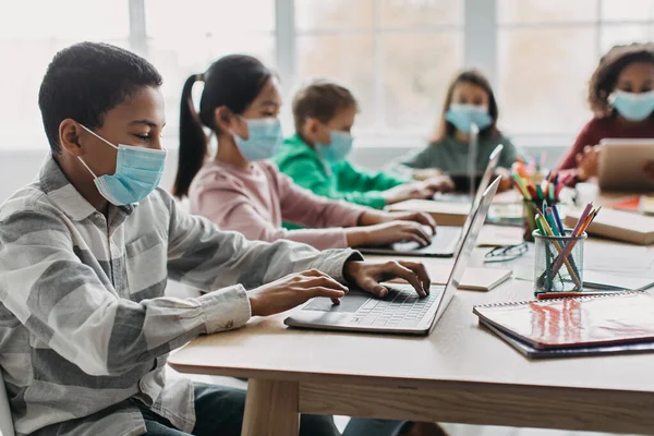 Online Education. Black Schoolboy Using Laptop Having Class With Multiracial Classmates Sitting At Desk In Modern Classroom Indoor, Wearing Face Masks. School And E-Learning. Selective Focus