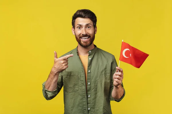 Happy man holding and pointing at China flag in his hand, posing and smiling at camera over yellow background. China citizenship, work and studying concept