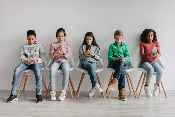 Group Of Amazed Diverse School Kids Using Smartphones Reading News And Browsing Internet Sitting In Chairs Over Gray Wall Indoors. Wow Application, Children And Gadgets Concept