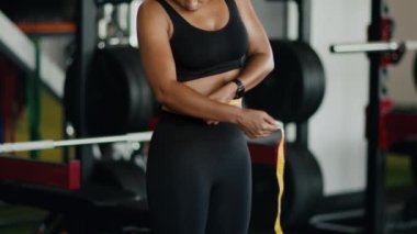 Slimming check. Unrecognizable black woman athlete measuring her waist and hips with tape, studying results of sport trainings at gym, slow motion, close up