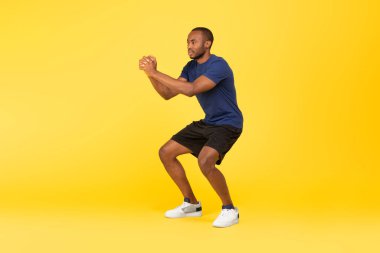 Athletic African American Man Doing Deep Squat Exercise Having Workout Over Yellow Studio Background, Wearing Sportswear. Healthy Fitness Concept. Full Length, Side View
