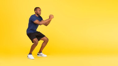 Sporty Black Guy Exercising Doing Deep Squats Training Looking Aside At Free Space For Text Over Yellow Studio Background. Fitness Workout Concept. Panorama, Full Length