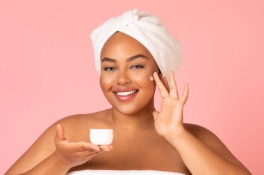 Smiling Black Female Applying Moisturizer On Face Enjoying Facial Skincare Routine Posing Over Pink Studio Background. Cosmetics For Skin And Beauty Care Concept