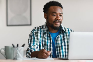 Black Man Freelancer Using Laptop Working And Taking Notes Sitting At Workplace In Modern Office. Web Technology And Remote Freelance Job Concept. Selective Focus