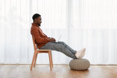 Side View Of Black Man Using Laptop Working Online Sitting In Chair Near Window At Home. Successful Freelancer Guy Websurfing Via Computer On Weekend. Technology Lifestyle Concept