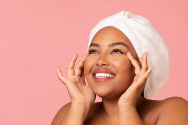 Facial Skincare. Happy African American Woman Moisturizing Face Skin Applying Moisturizer Posing With Wrapped Towel On Head Looking Aside Over Pink Background. Studio Shot